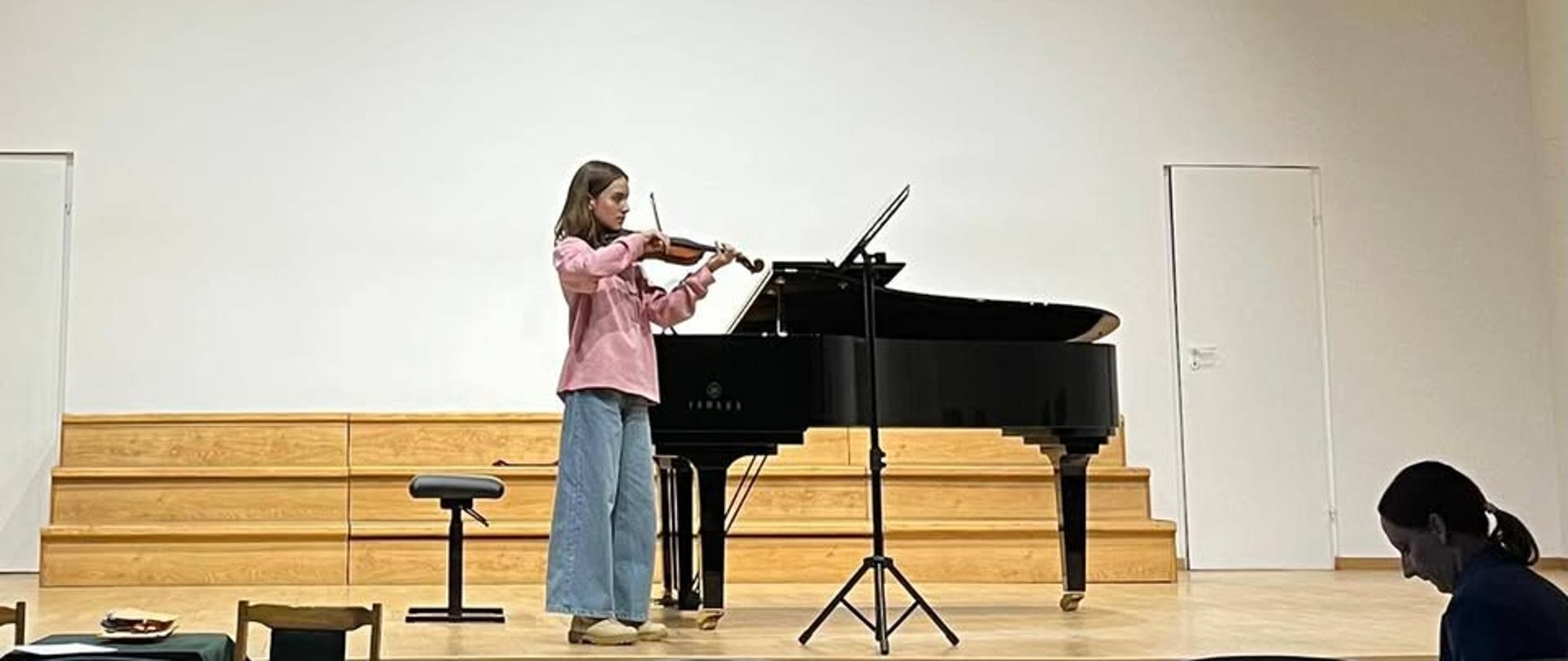 Młoda dziewczyna w różowej koszuli i szerokich jeansach gra na skrzypcach na oświetlonej scenie sali koncertowej. Stoi obok otwartego czarnego fortepianu marki Yamaha. W tle widać białą ścianę, drewniane podesty oraz nowoczesne oświetlenie sufitowe. Widok z perspektywy widowni, z rzędami ciemnych foteli w dolnej części kadru.