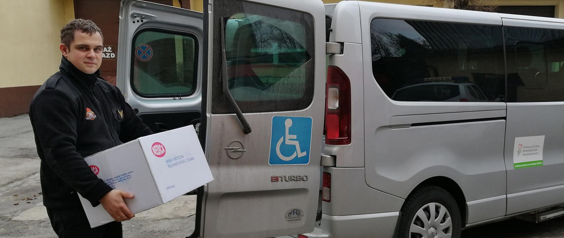 Strażak PSP stoi za samochodem dostawczym i trzyma w rękach paczkę z maseczkami ochronnymi.