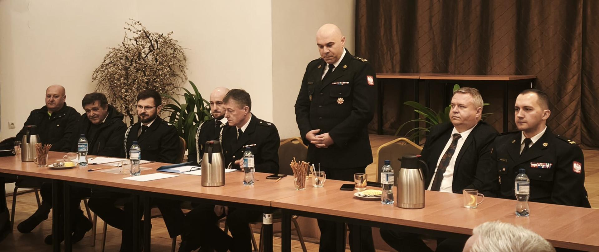 Zdjęcie przedstawia zastępce podkarpackiego komendanta wojewódzkiego podczas zebrania OSP. Obok siedzi burmistrz Ustrzyk Dolnych i zastępca komendanta powiatowego. Po drugiej stronie druhowie OSP. 