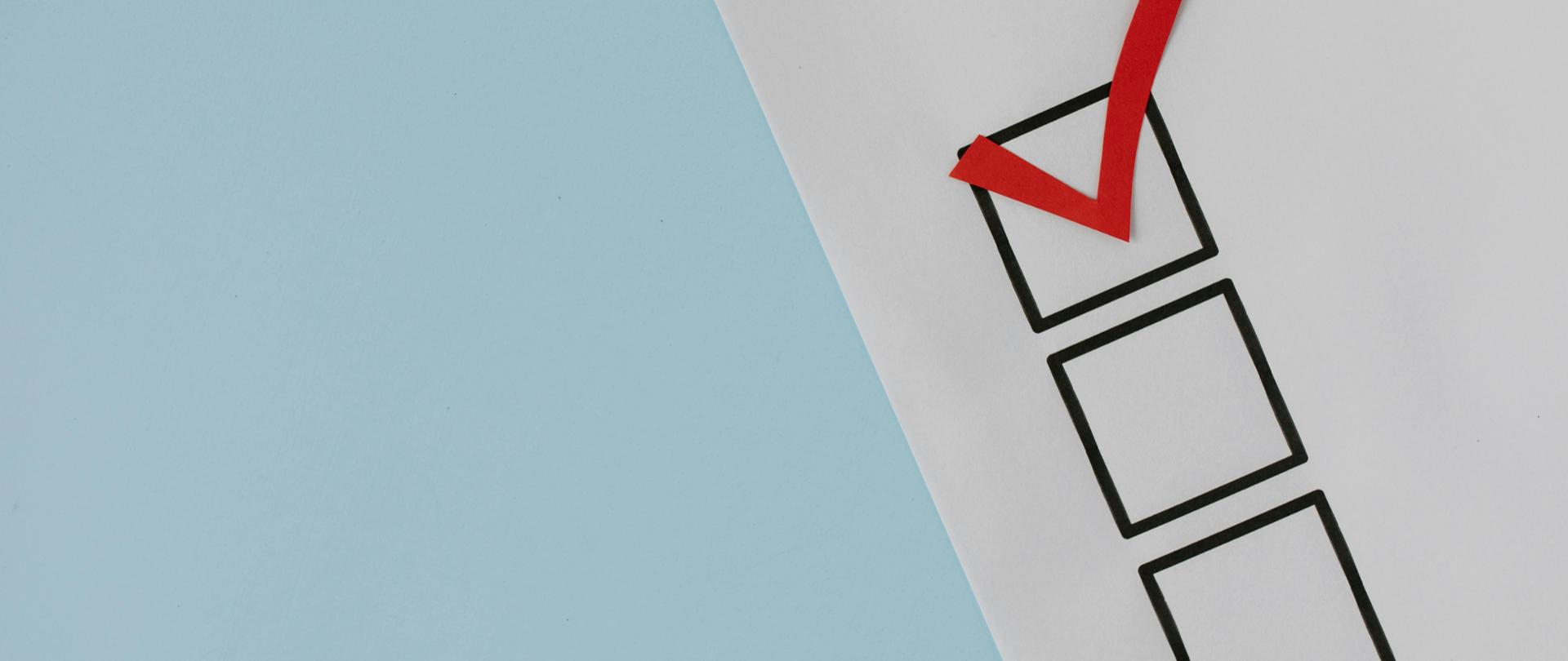 na jasnoniebieskim tle kawałek białej kartki na której znajdują się trzy czarne kwadraty w pozycji pionowej a w pierwszym od góry znajduje się zaznaczenie tak zwany ptaszek w kolorze czerwonym