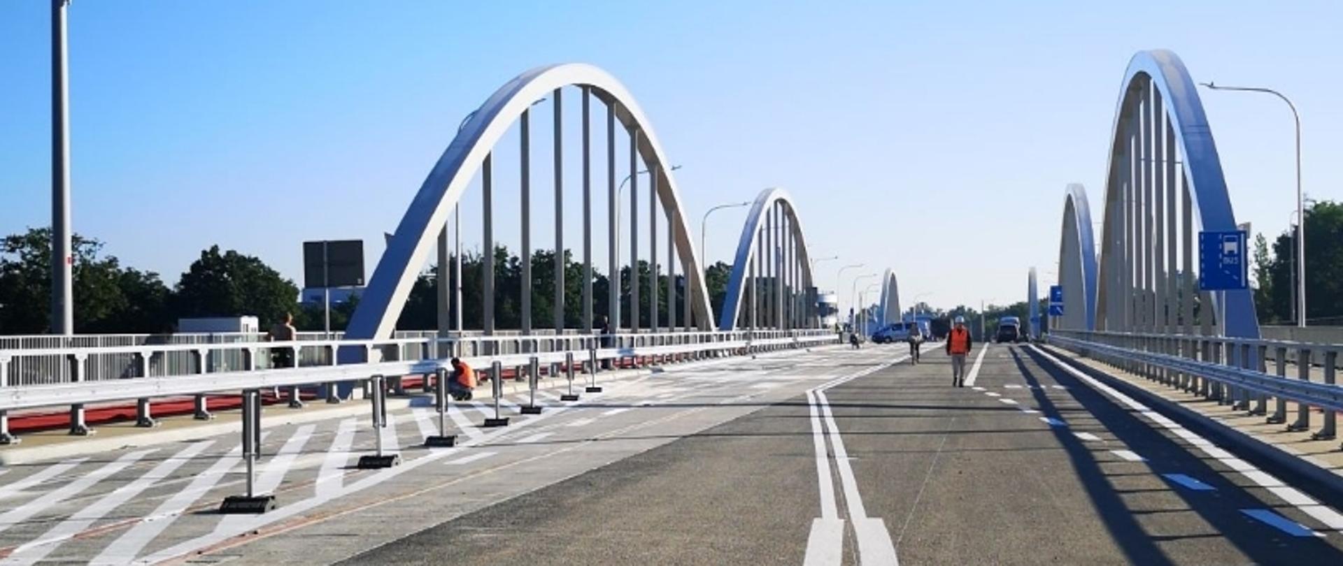 Most z białymi łukami, pasami ruchu, chodnikami i latarniami; ludzie spacerują w słoneczny dzień.