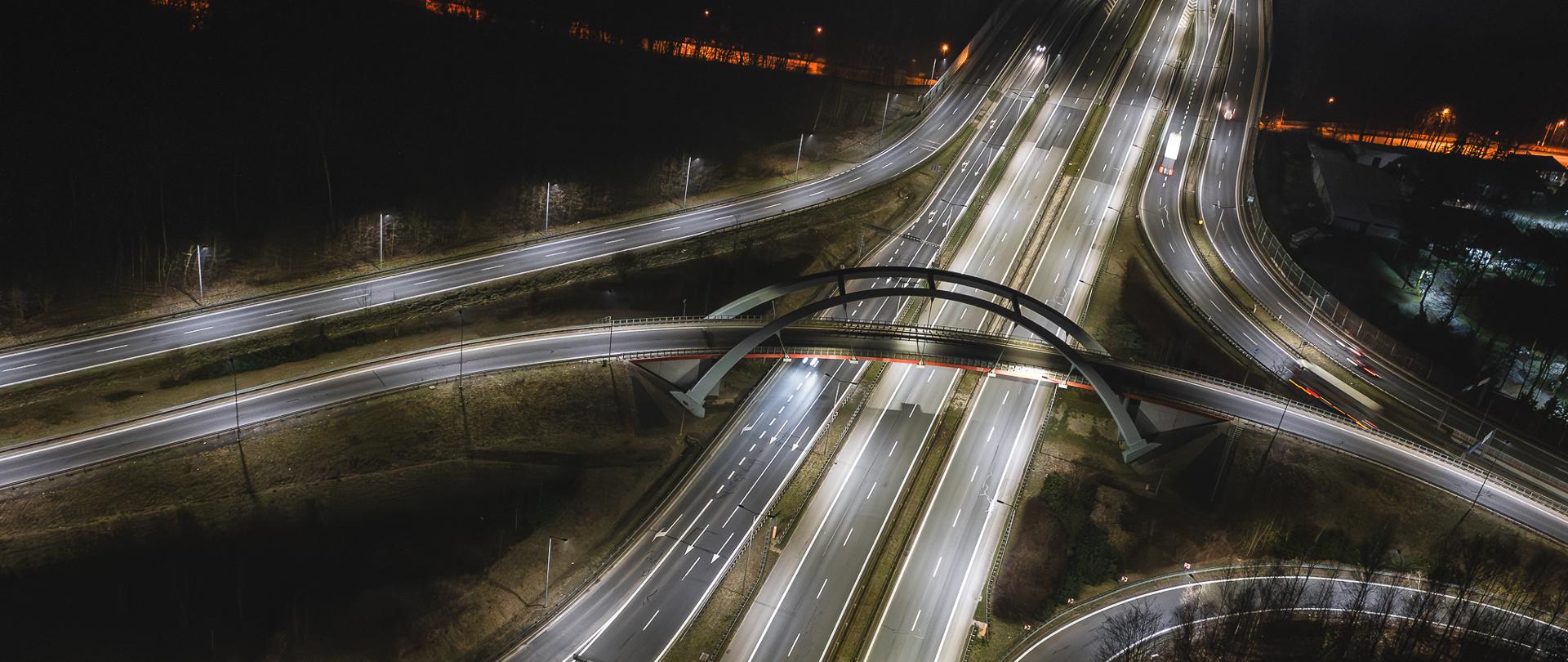 Fotografia przedstawia węzeł Murckowska w Katowicach, uchwycony nocą z perspektywy lotu ptaka. Nowoczesne ledowe oświetlenie efektownie podkreśla strukturę dróg, tworząc dynamiczną kompozycję świetlnych linii przecinających ciemne otoczenie. Jasne światła LED poprawiają widoczność i bezpieczeństwo kierowców, uwydatniając wielopasmowe autostrady oraz estakady przecinające przestrzeń miejską. W tle widać otaczającą infrastrukturę, w tym latarnie uliczne i fragmenty zieleni. Cała scena oddaje modernizację i wysoką jakość technologii zastosowanej w tym ważnym punkcie komunikacyjnym miasta.