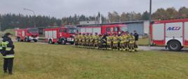 Zakończenie ćwiczeń na tle strażackich samochodów stoją w dwuszeregu strażacy w piaskowych mundurach.