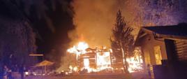 POŻAR LOKALU GASTRONOMICZNEGO. Na zdjęciu pożar drewnianego budynku, ogniem objęty jest cały budynek, obok zabudowania i drzewa 