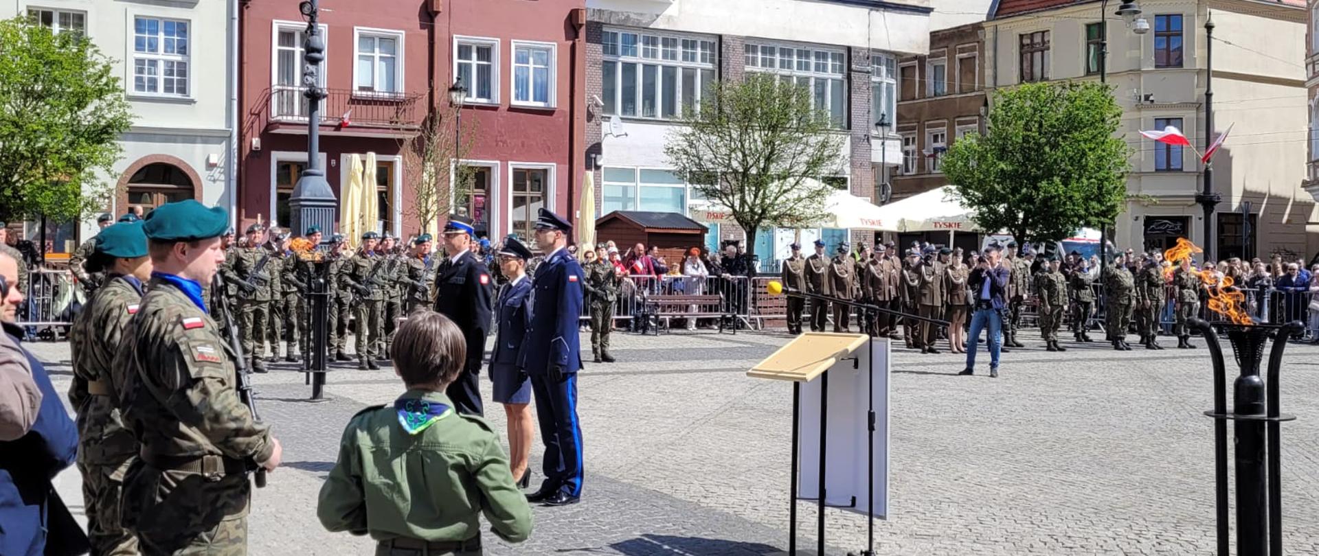 Trzej funkcjonariusze służb mundurowych w trakcie składania wiązanki na uroczystości patriotycznej. W tle pododdziały Wojska Polskiego i zebrany tłum.
