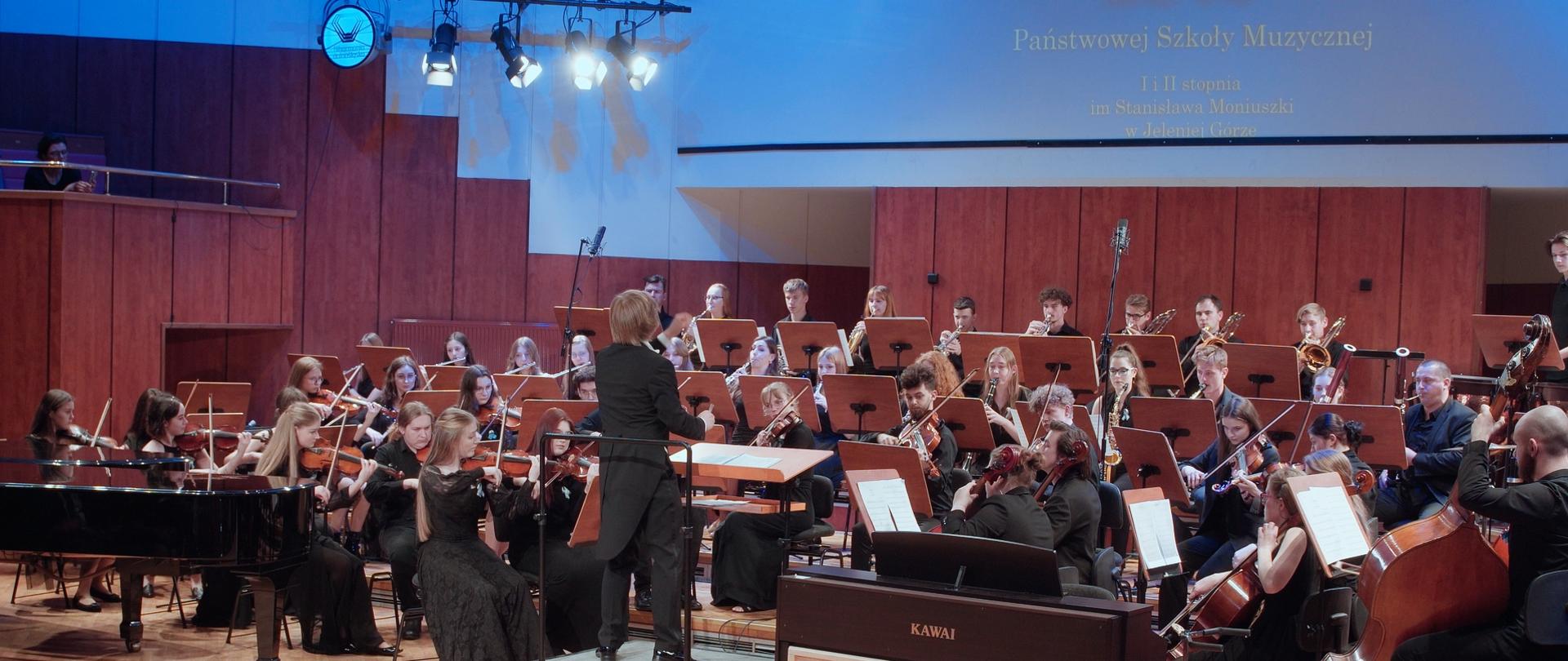 Zdjęcie przedstawiające kadr z koncertu, w filharmonii dolnośląskiej, szkolnej orkiestry symfonicznej PSM II stopnia. Na zdjęciu widoczny cały skład orkiestry wraz z dyrygentem