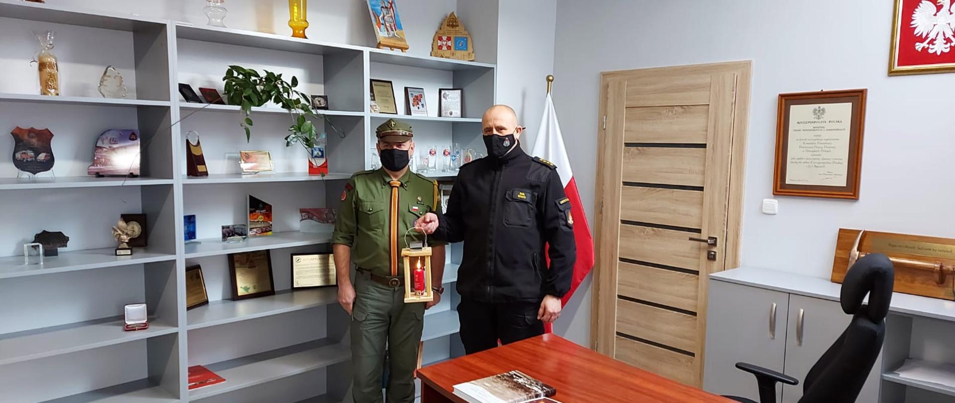 Na zdjęciu widać komendanta powiatowego PSP w Ustrzykach Dolnych ubrany w polar który trzyma w ręku zapalony lampion. Obok komendanta stoi harcerz w zielonej koszuli oraz czapce, szarych spodniach i złotym krawacie. Zdjęcie wykonane w biurze komendanta, po prawej widać biurko, a za obecnymi na zdjęciu flaga państwowa i półki na których stoją głównie dyplomy.
