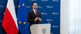 Minister Jakub Jaworowski stoi za mównicą. W tle ścianka z logotypami MAP i flagi. 