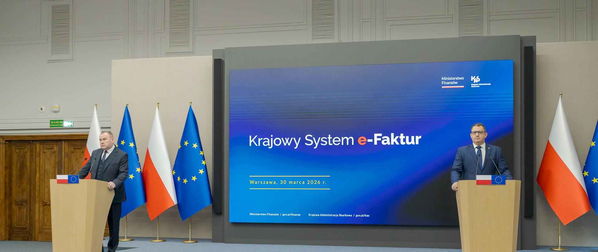 Marcin Łoboda szef Krajowej Administracji Skarbowej oraz Zbigniew Stawicki zastępca szefa KAS podczas konferencji prasowej w Ministerstwie Finansów opowiedzieli o wdrożeniu II etapu Krajowego Systemu e-Faktur przy mównicach przed dużym ekranem z napisem „Krajowy System e‑Faktur”, obok flag Polski i Unii Europejskiej