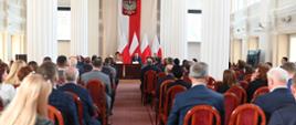 Widok z tyłu na salę wypełnioną ludźmi siedzącymi na krzesłach, przed nimi na tle polskich flag siedzi za stołem minister Czarnek i kobieta w jasnym ubraniu.