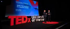 Zdjęcie z boku, w zaciemnionej sali na podwyższeniu stoi wiceminister Gzik i mężczyzna w garniturze, za nimi napis TEDx University of Warsaw ułożony z dużych liter, nad nim wielki ekran.