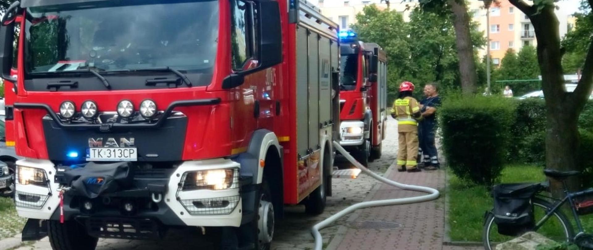 Zdjęcie przedstawia dwa samochody pożarnicze, które stoją na drodze osiedlowej z podpiętym wężem gaśniczym. 