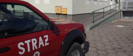 W lewym dolnym rogu widać prawy przód auta strażackiego osobowego koloru czerwonego z białym napisem na drzwiach Straż, w tle budynek przychodni, w której realizowane są szczepienia. 
