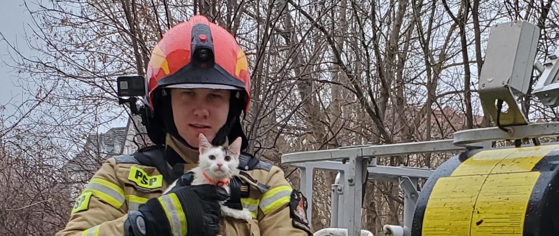 Strażak soi przy koszu podnośnika i na rękach trzyma uratowanego biało czarnego kotka. 