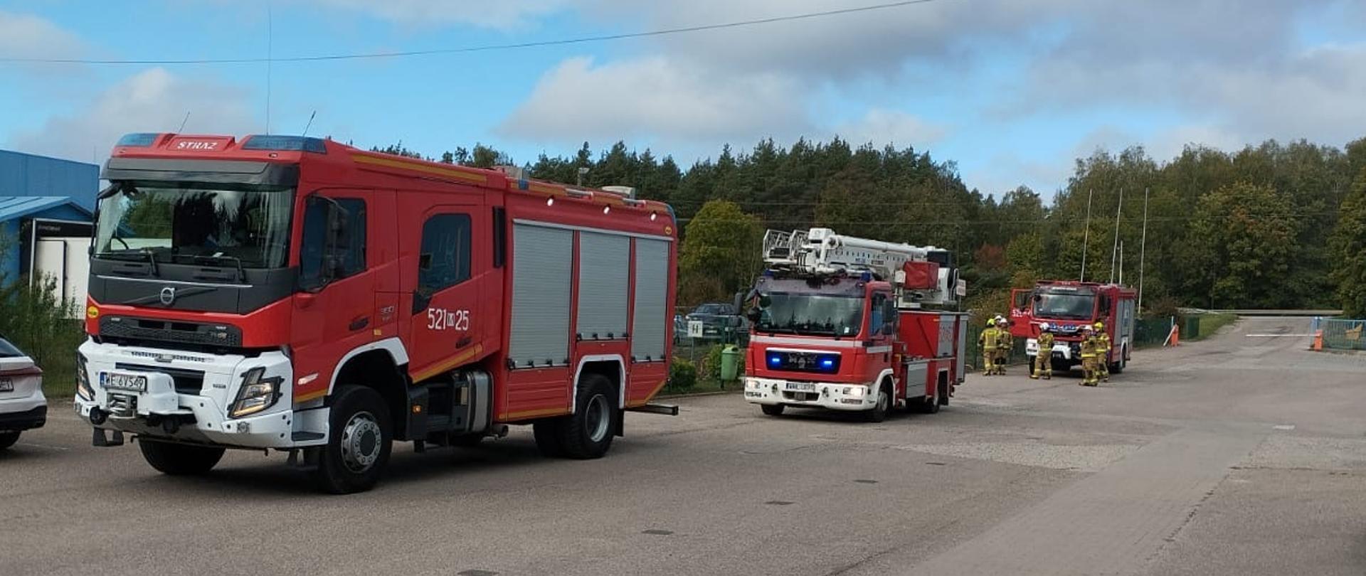 Na szerokiej asfaltowej drodze zaparkowane są trzy wozy strażackie. Najbliżej znajduje się czerwony pojazd ratowniczo-gaśniczy, za nim wóz z drabiną mechaniczną, a dalej kolejny pojazd strażacki. Na miejscu obecni są strażacy przygotowujący się do akcji. W tle widoczna jest zieleń – drzewa i krzewy – oraz niebo z częściowym zachmurzeniem.
