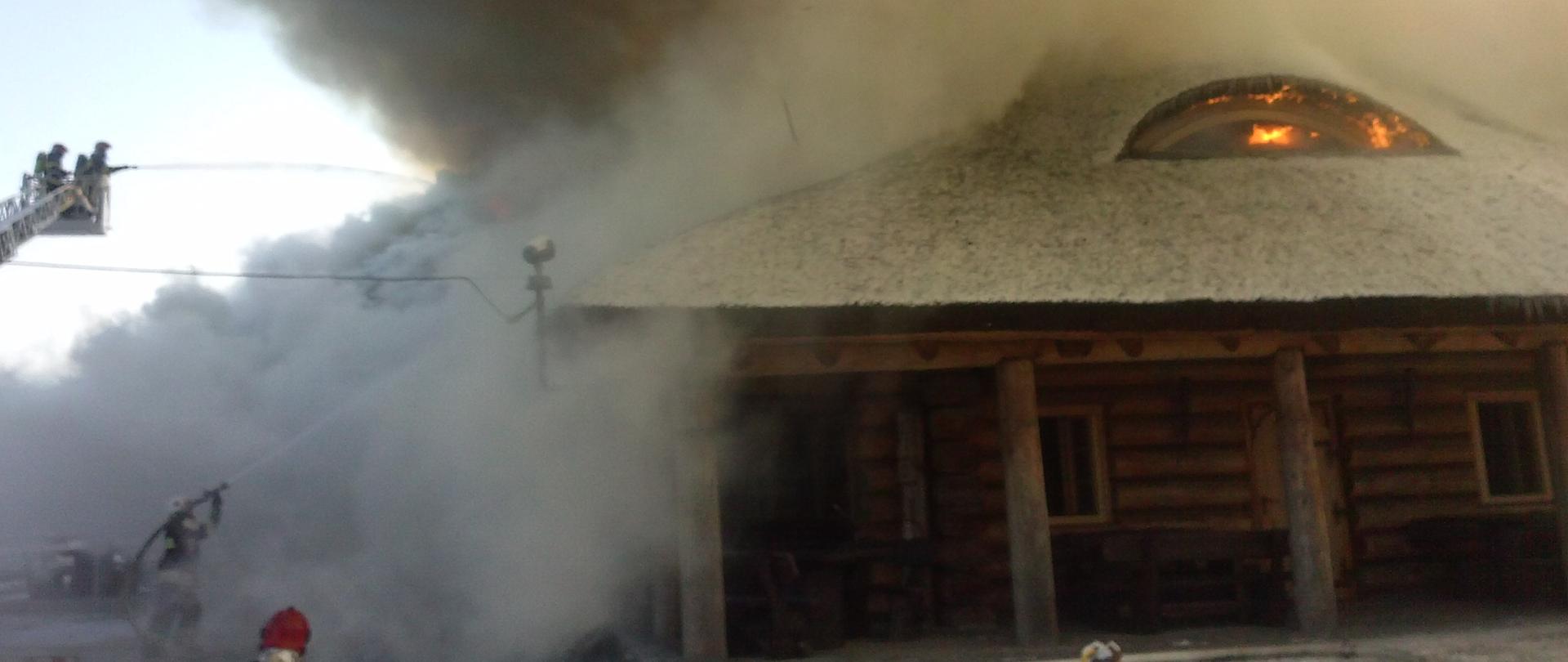 płonący dach drewnianego, parterowego budynku, po lewej stronie strażacy na podnośniku gaszący pożar