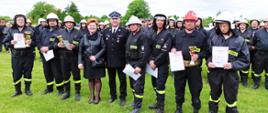 Zdjęcie przedstawia 8 strażaków OSP ustawionych w szeregu. Ubrani są w ubranie koszarowe, pasy strażackie oraz hełmy. W dłoniach trzymają dyplomy i puchary. Po między nimi stoi kobieta (wójtowa). Za nimi stoją pozostali druhowie w dwuszeregu.
