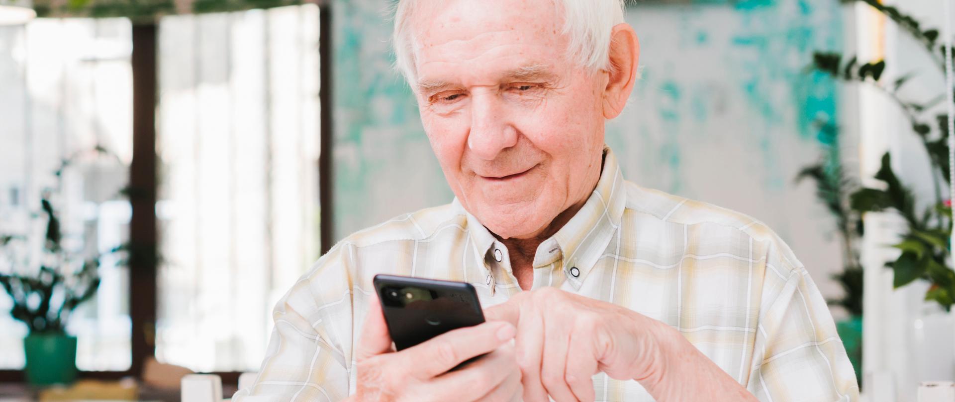 Zdjęcie ilustracyjne w poziomie. Na pierwszym planie starszy mężczyzna, ubrany w białą koszulę, siedzi przy stoliku. W ręku trzyma smartfona. 