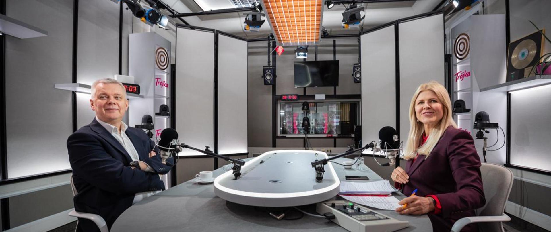 Na zdjęciu studio radiowe. Na pierwszym planie okrągły stół radiowy z zamontowanymi mikrofonami. Po lewej stronie stołu, za mikrofonem siedzi minister Tomasz Siemoniak, natomiast po prawej stronie, również za mikrofonem - redaktor Renata Grochal. Na stole znajdują się filiżanki, notatki, telefon, mikser radiowy. Pod sufitem znajdują się lampy. W tle zdjęcia widać również szybę, ekran, kolumny. Po prawej i lewej stronie zdjęcia białe szafki z różowymi napisami "Trójka".