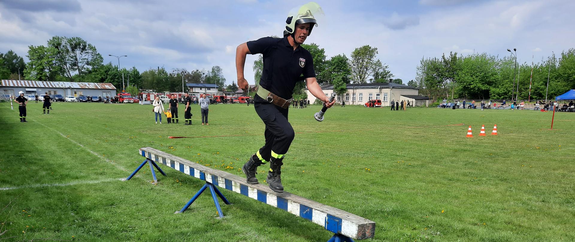 Strażak biegnący po równoważni w pasie, białym hełmie i z prądownicą w ręku.