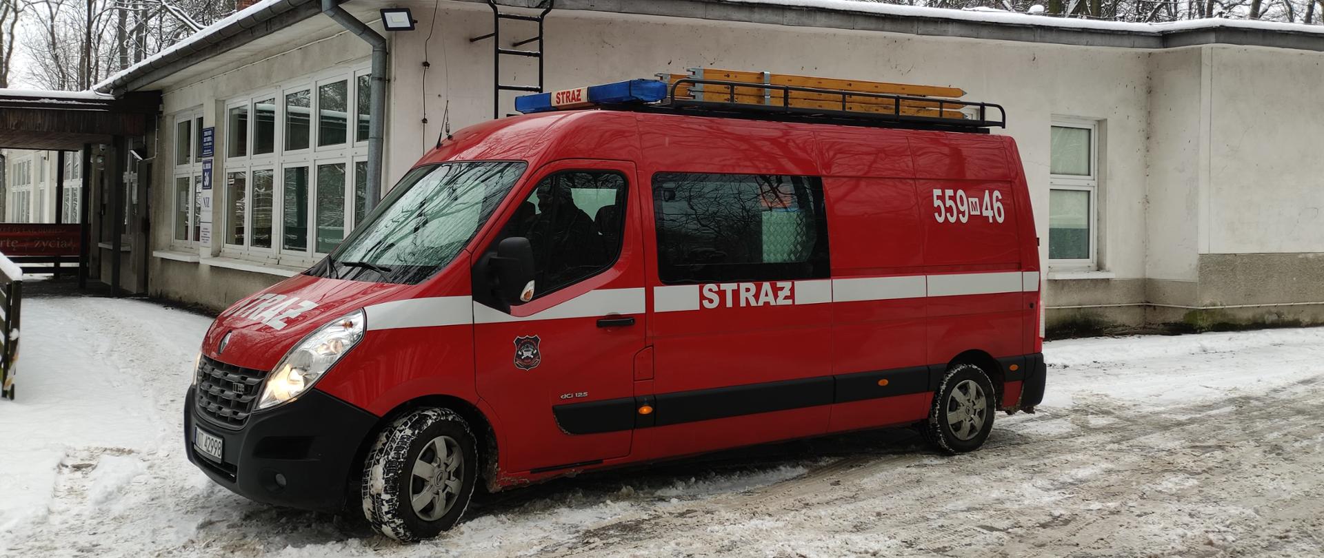 Zdjęcie przedstawia samochód strażacki, którym realizowany jest transport seniorów do punktów szczepień przeciwko COVID 19. W tle znajduję się przychodnia