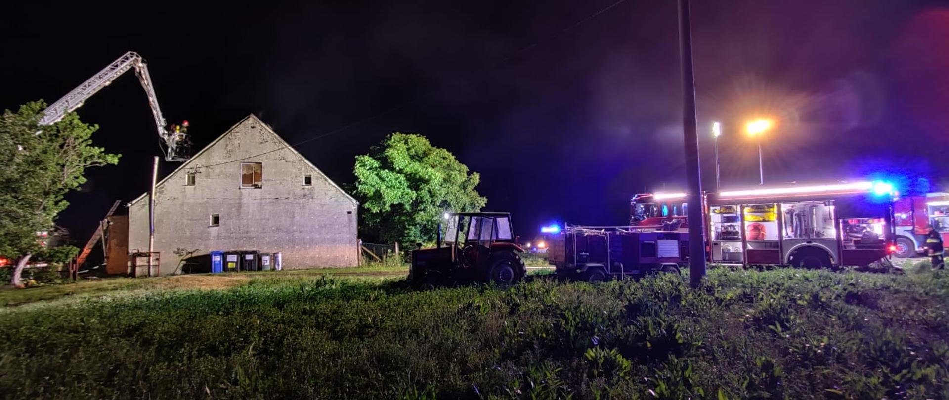 Pora nocna, sztuczne oświetlenie. Na pierwszym planie kontury ciągnika rolniczego, w oddali oświetlony szczyt budynku mieszkalnego a przy nim rozłożone ramię podnośnika strażackiego. W prawej stronie kadru samochody strażackie w złączonymi światłami błyskowymi w kolorze niebieskim.
