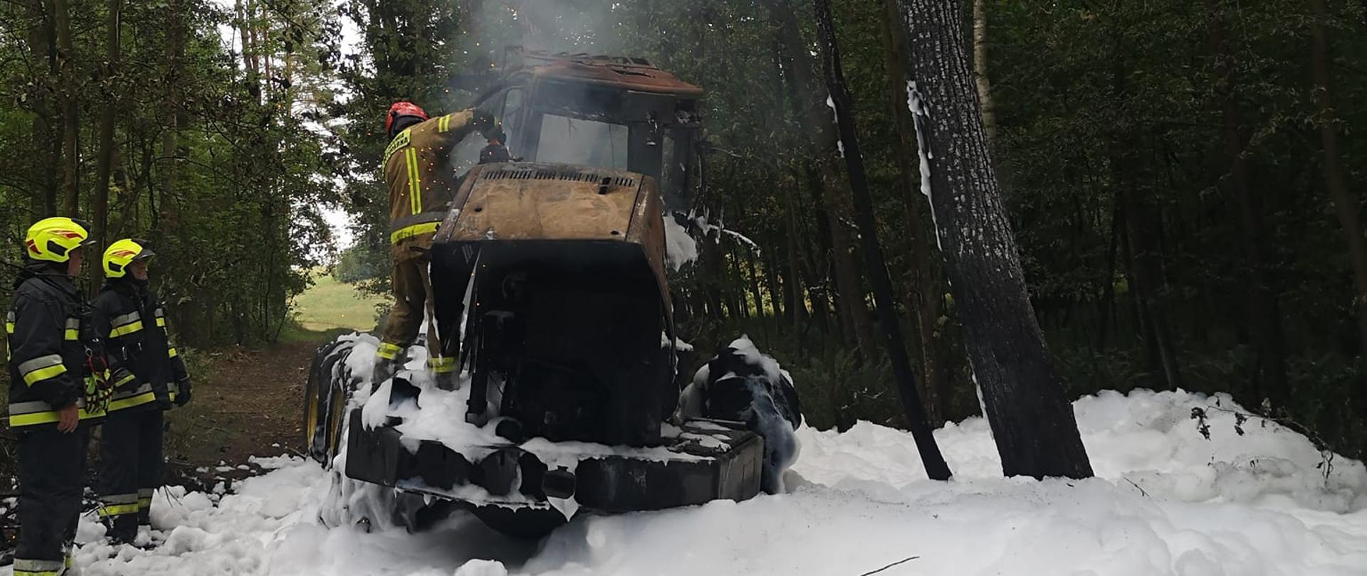 Zdjęcie przedstawia spaloną maszynę leśną typu Harwester pokrytą piana gaśniczą. strażak stoi na maszynie i wykonuje pracę rozbiurkowe maszyny.
