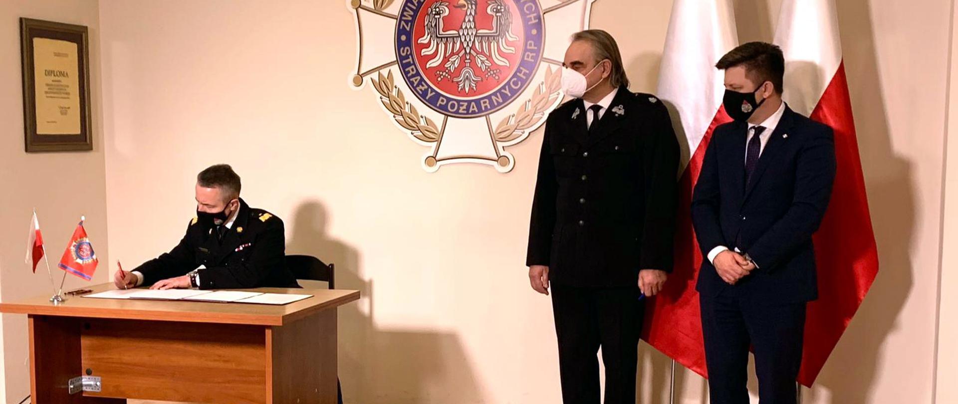 Uczestnicy spotkania podpisują list intencyjny. W tle znak OSP i flagi Polski. W trakcie podpisu Komendant Główny PSP nadbryg. Andrzej Bartkowiak.