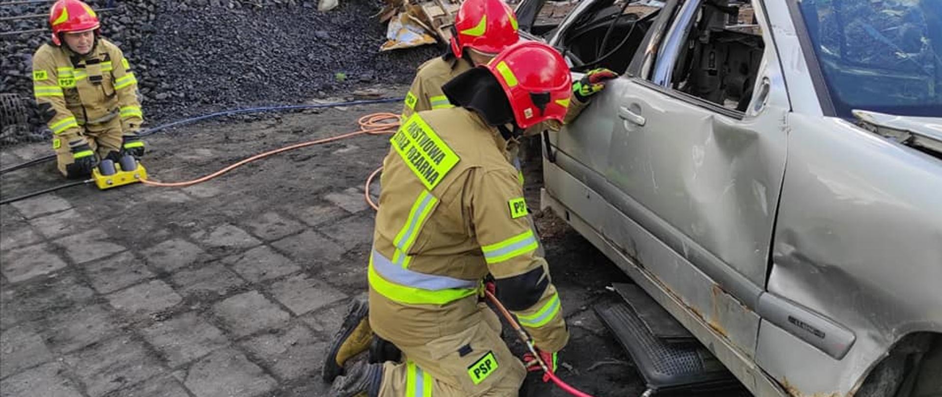 Na zdjęciu widać trzech strażaków ratowników ubranych w ubrania specjalne i hełmy, którzy przy pomocy sprzętu pneumatycznego unoszą prawy bok pojazdu. W tle widać złom i węgiel.