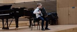 Uczeń siedzi na krześle i gra na akordeonie na scenie sali koncertowej PSM, za nim stoi fortepian