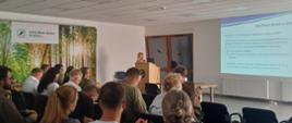 Spotkanie w sali konferencyjnej. Przy na krzesłach ustawionych w rzędach siedzą uczestnicy, a przy mównicy prezentację prowadzi kobieta. Na ekranie wyświetlana jest prezentacja dotycząca weryfikacji danych w urzędowym rejestrze podmiotów profesjonalnych. W tle widoczna jest grafika lasu z napisem „Leśny Bank Genów Kostrzyca”.