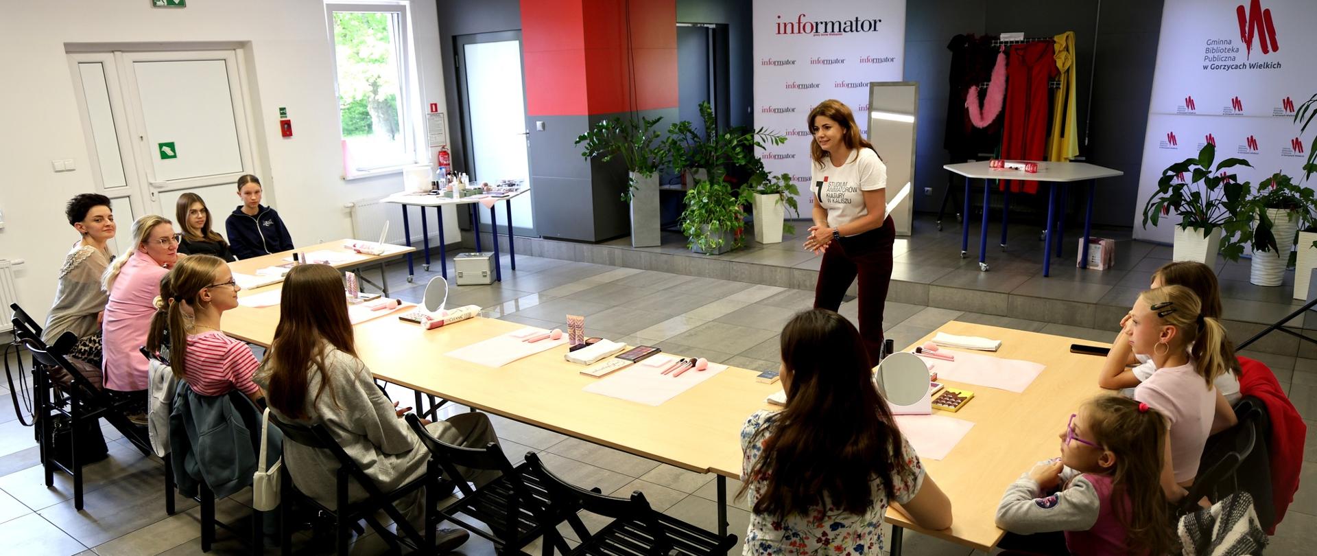 Warsztaty z charakteryzacji prowadzone w sali Gminnej Biblioteki Publicznej w Gorzycach Wielkich. Przy stołach siedzą dzieci i młodzież, a prowadząca ze Studium Animatorów Kultury w Kaliszu omawia przygotowane materiały do pracy.