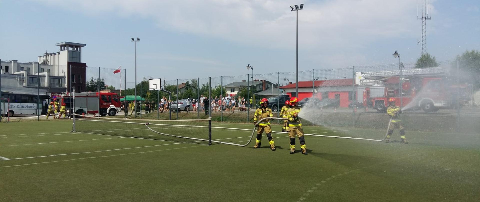 Na pierwszym planie czterech strażaków w ubraniach bojowych z rozwiniętymi dwiema liniami gaśniczymi leją wodę. W oddali na tle strażnicy wozy strażackie oraz odwiedzający ludzie.