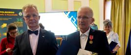 W sali z żółto-białymi ścianami stoi obok siebie dwóch mężczyzn w garniturach, jeden ma przypięty do marynarki medal i trzyma w ręku dyplom.