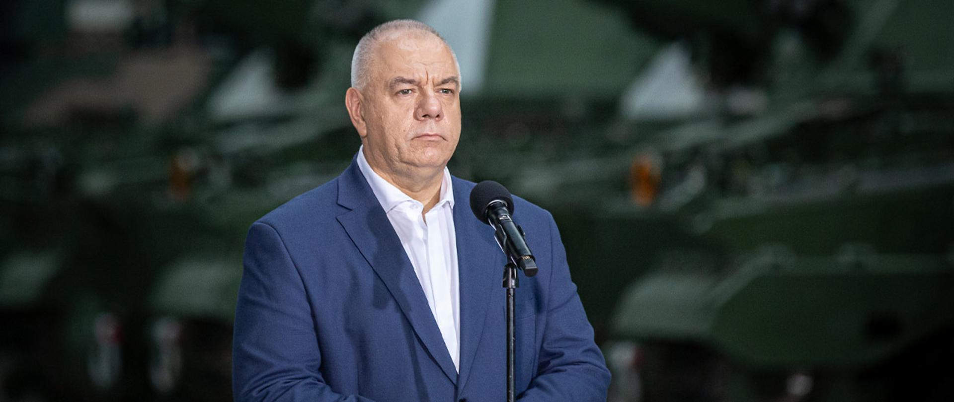 Wicepremier Jacek Sasin stoi za mikrofonem. W tle czołgi. 