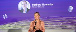 Minister Edukacji Barbara Nowacka stojąca na scenie w pudroworóżowej sukience, przemawiająca z mikrofonem w ręku. W tle znajduje się wielki fioletowy ekran z jej portretem, imieniem i nazwiskiem oraz funkcją. Na pierwszym planie widać sylwetki widowni.