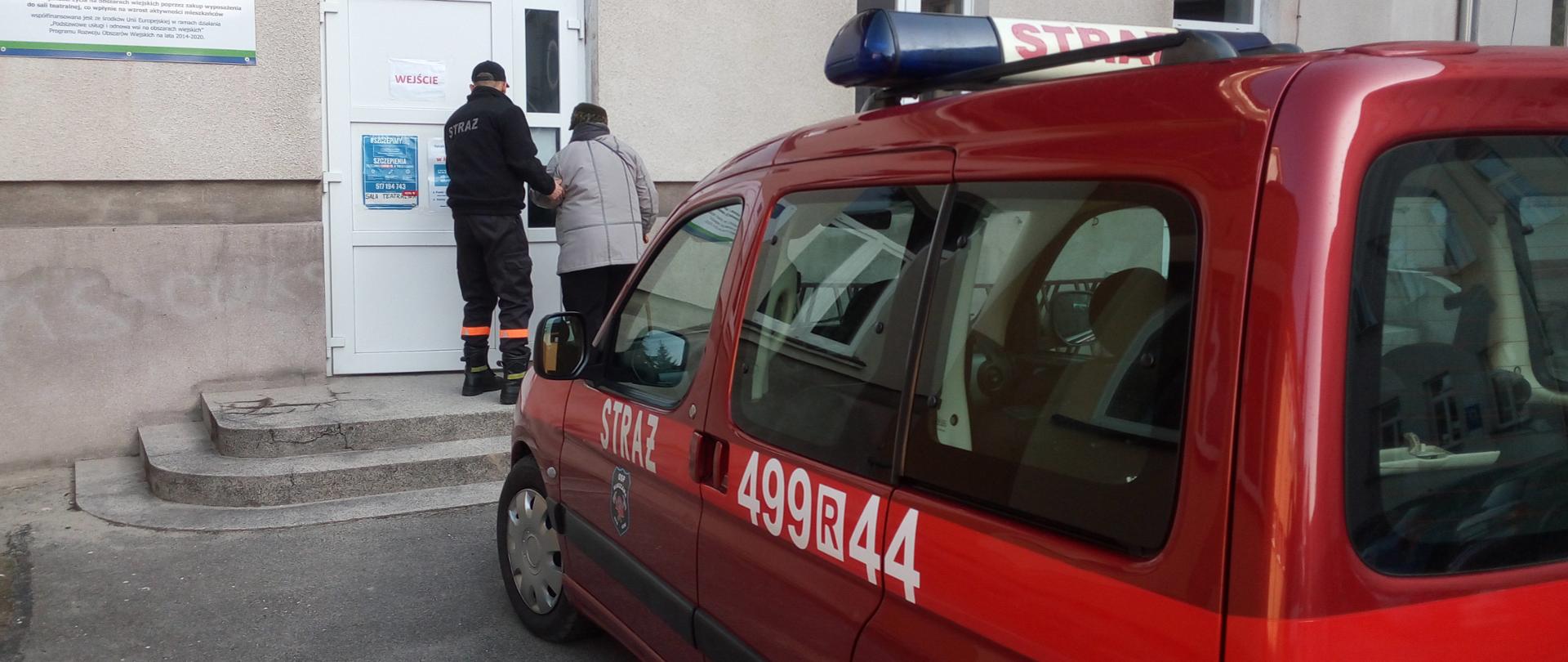 Zdjęcie przedstawia plac na którym zlokalizowany jest punkt szczepień w miejscowości Rakszawa. Na pierwszym planie widoczny jest samochód operacyjny Ochotniczej Straży Pożarnej w Rakszawie. Na drugim planie strażak tej jednostce pomaga starszej osobie o ograniczonej zdolności poruszania się w wejściu do budynku punktu szczepień.