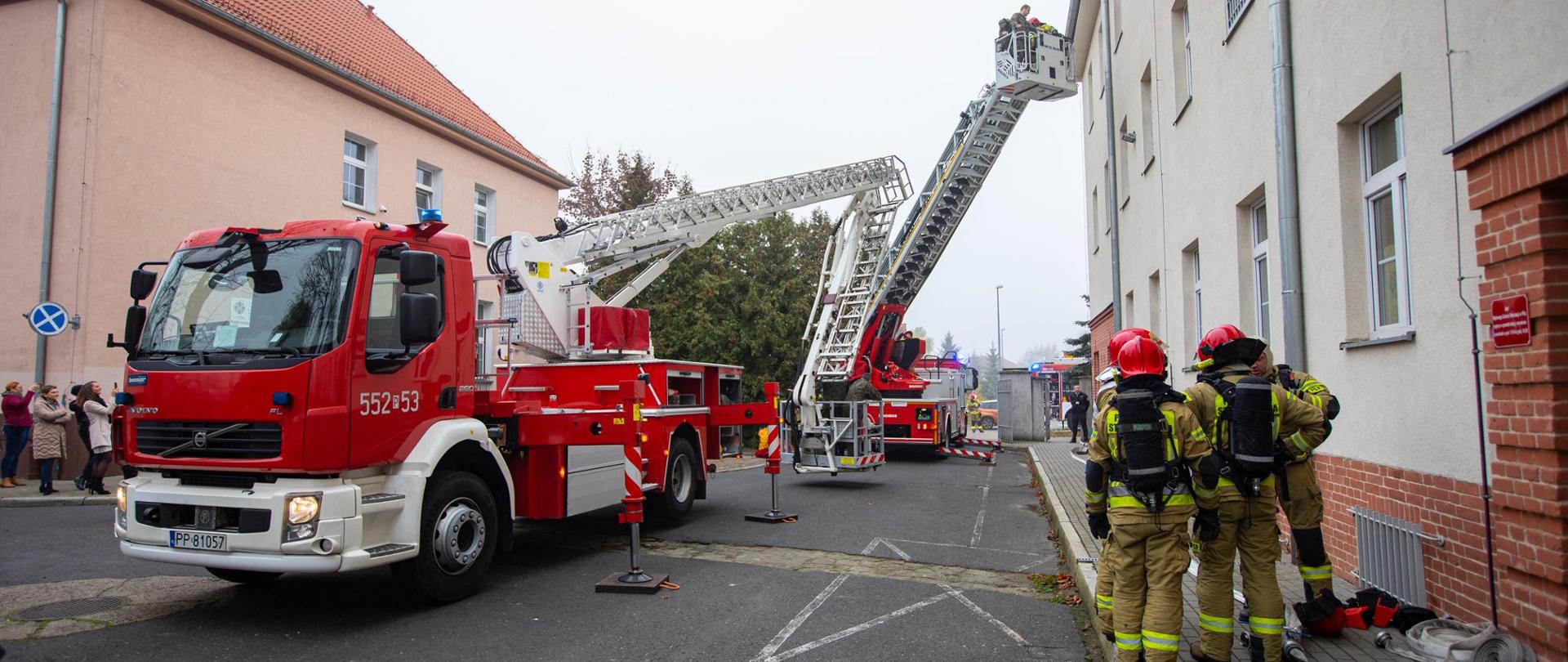 Na zdjęciu strażacy przygotowują się do wejścia w strefę niebezpieczną. w Tle widać podnośnik hydrauliczny oraz drabinę mechaniczną.