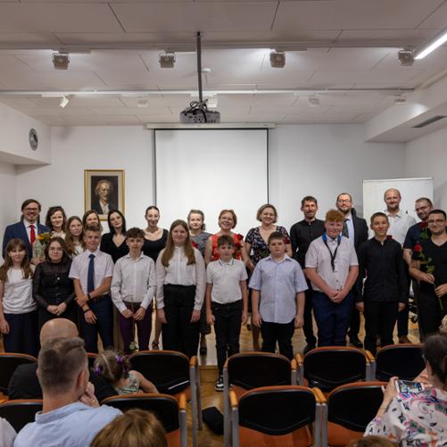 Zdjęcie grupowe przedstawiające absolwentów roku szkolnego 2022/2023 wraz z ich nauczycielami. Nauczyciele stoją na scenie PSM w Głubczycach, absolwenci przed stopniem stanowiącym granicę sceny. Na dole zdjęcia widać również kilka osób siedzących na publiczności.