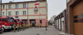 Strażacy stoją w dwóch rzędach na przeciwko siebie, uroczyście podnosząc flagę Rzeczypospolitej Polskiej na maszt.