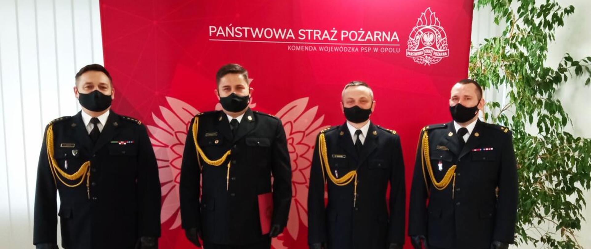 Zdjęcie przedstawia czterech strażaków w rzędzie w umundurowaniu galowym, twarzą skierowanych w kierunku fotografującego. Zdjęcie wykonane podczas uroczystości wręczenia awansu na pierwszy stopień oficerski PSP - młodszego kapitana - dla sekc. Ryszarda Kowalczyka funkcjonariusza KW PSP Opole. Od lewej stoją: Opolski Komendant Wojewódzki PSP st. bryg. Krzysztof Kędryk, mł. kpt. Ryszard Kowalczyk awansowany, Zastępca Opolskiego Komendanta Wojewódzkiego PSP bryg. Wojciech Nawara oraz mł. bryg. Grzegorz Brzozowski Naczelnik Wydziału Kwatermistrzowskiego.
W na drugim planie plansza koloru czerwonego z białym napisem po jej prawej stronie "Państwowa Straż Pożarna" a poniżej "Komenda Wojewódzka PSP w Opolu". Po prawej stronie znajduje się białe logo PSP a w 2/3 wielkości planszy po jej lewej stronie znajduje się biały orzeł (godło). Po prawej stronie planszy znajduje się roślina z liśćmi koloru zielonego wysokości planszy. Wszyscy strażacy mają założone czarne maseczki na części twarzy.