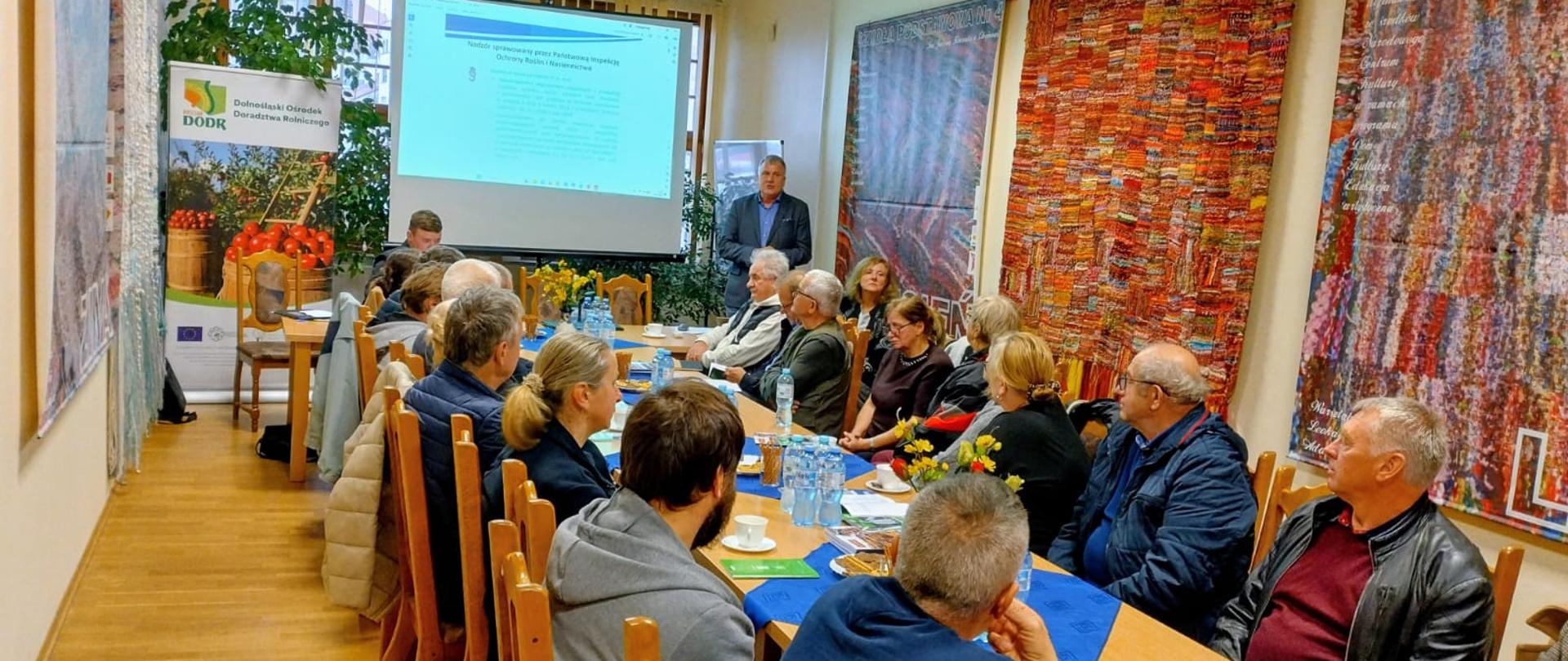 Rolnicy siedzą przy podłużnym stole, w tle widoczny ekran z prezentacją, przy którym po prawej stronie stoi wykładowca