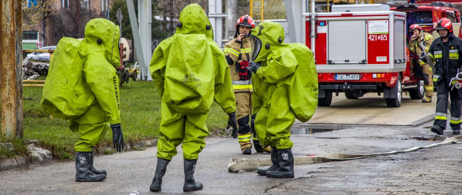 Strażacy ze SGRChem "Kędzierzyn-Koźle" przygotowują się do wejścia w strefę niebezpieczną. Strażacy w specjalnym ubraniu chemicznym przygotowani do realizacji zadań w ciężkim terenie. W tle widoczni inni strażacy PSP oraz pojazdy grupy specjalistycznej. Ćwiczenia pk. "Silekol 2023", które odbyły się 19 kwietnia 2023 roku w Kędzierzynie-Koźlu.