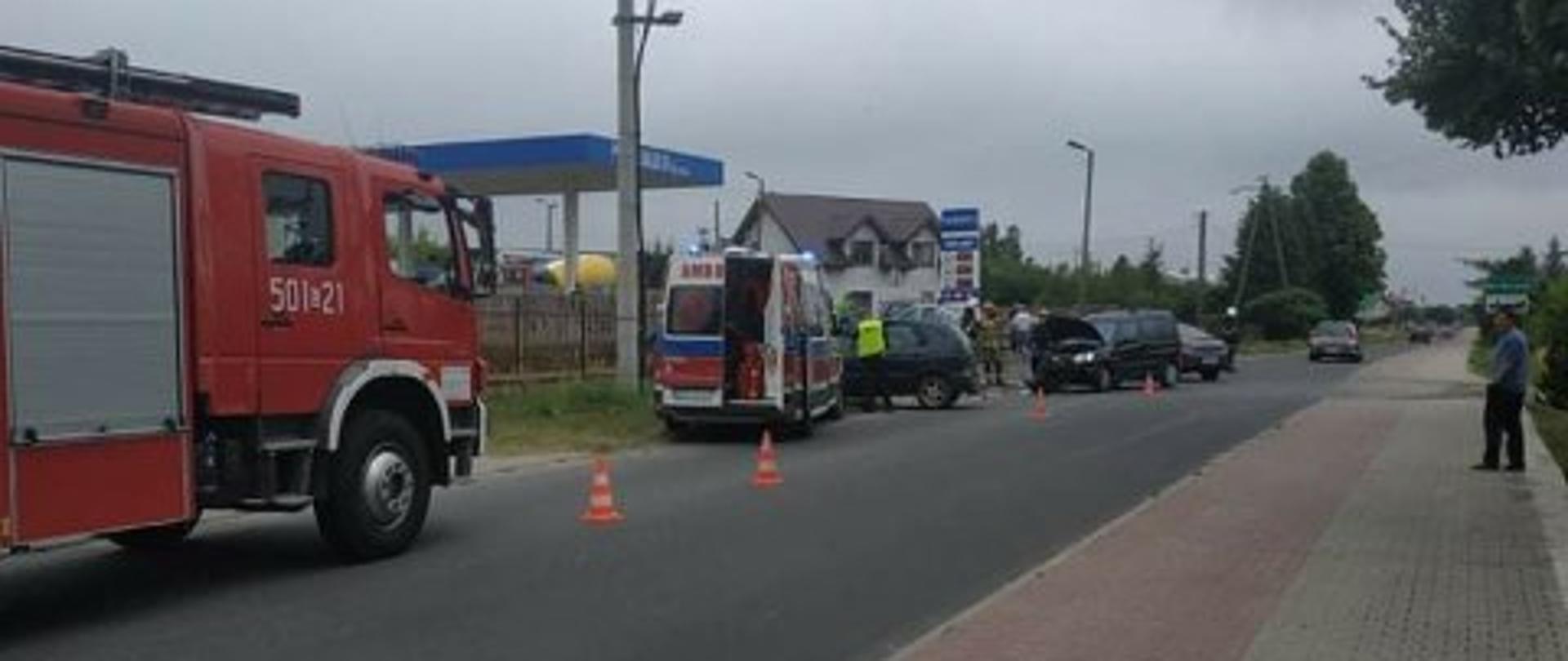 Na ulicy po lewej stronie stoi wóz strażacki, przed nik karetka, a przed nią dwa samochody osobowe,. Widać że są po zderzeniu, w jednym uniesiona jest do góry przednia maska 