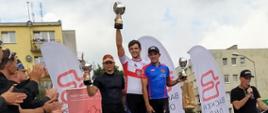 Zdjęcie przedstawia trzech strażaków ubranych w sportowe stroje kolarskie, którzy stoją na podium. Strażacy trzymają puchary i wznoszą je do góry. Po lewej stronie zdjęcia znajdują się osoby, które biją brawo zawodnikom. W tle widnieją budynki mieszkalne