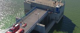 Zdjęcie wykonane podczas ćwiczeń zgrywających współdziałanie różnych podmiotów dbających o bezpieczeństwo publiczne. Ćwiczenia zorganizowane na zalewie wodnym w miejscowości Domaniów. Na zdjęciu widać fragment zapory wodnej. Zapora zbudowana jest z betonu. Na drodze technicznej będącej częścią zapory widać ustawione dwa pojazdy terenowe typu pick-up należące do Państwowej Straży Pożarnej. Na zaporze stoją strażacy ze specjalistycznej grupy ratownictwa wysokościowego. Ratownicy wykonują pozorowaną ewakuację osoby poszkodowanej która spadła poniżej zapory. Do tego celu używają lin alpinistycznych oraz noszy. Zdjęcie wykonane w formie widoku z góry przy użyciu drona.