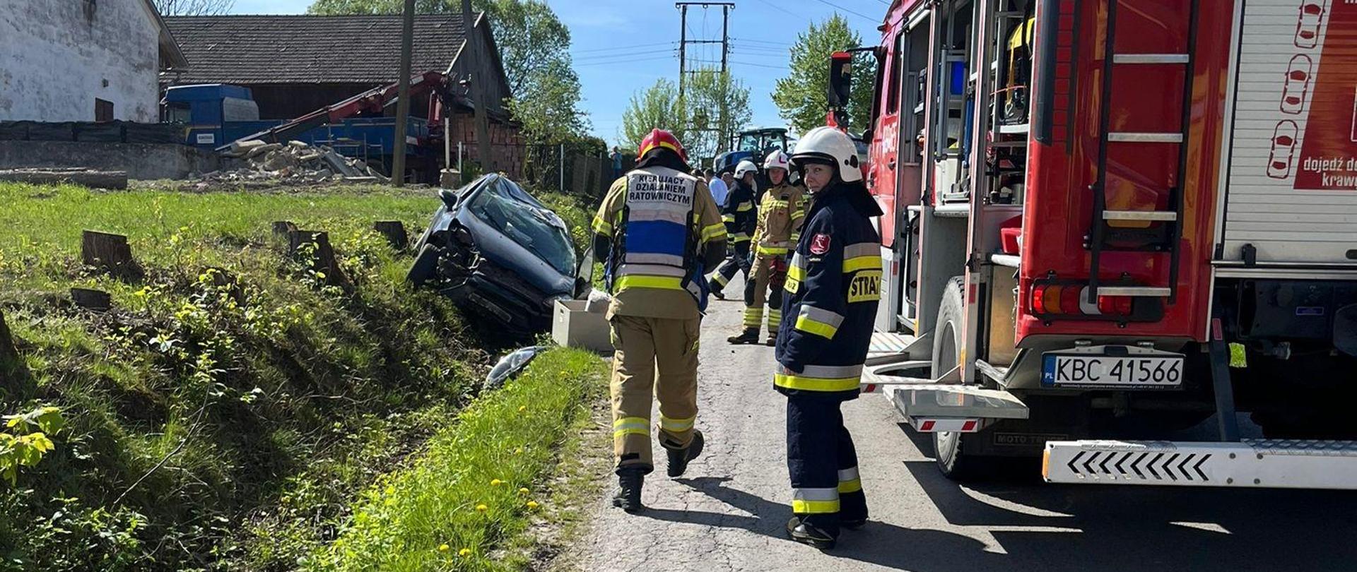 Zdjęcie przedstawia dachowanie pojazdu osobowego. Na zdjęciu widać uszkodzony samochód osobowy który leży na boku w rowie. Obok pojazdu stoją strażacy ubrani w ubrania specjalne i hełmy. Jeden z nich ma założoną kamizelkę z napisem kierujący działaniami ratowniczymi. Obok strażaków stoi czerwony pojazd pożarniczy który ma otwarte skrytki w których znajduje się sprzęt pożarniczo-ratowniczy. Po lewej stronie widać budynki.