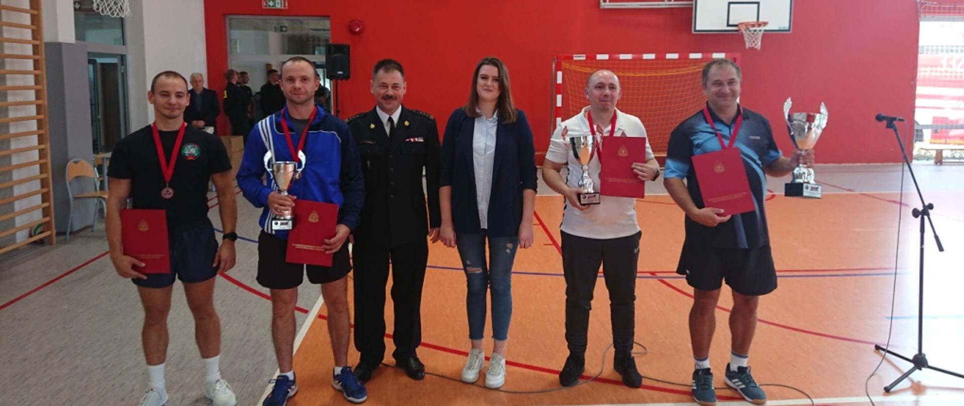 Zdjęcie wykonane podczas rozgrywek finału mistrzostw województwa mazowieckiego strażaków w tenisie stołowym. Zdjęcie wykonane w pomieszczeniu Sali gimnastycznej gdzie przeprowadzono rozgrywki. Na zdjęciu widać ustawionych w rzędzie pozujących do pamiątkowej fotografii zawodników którzy zajęli czołowe miejsca w obecności zastępcy komendanta miejskiego Państwowej Straży Pożarnej w Radomiu – starszego brygadier Marcina Ziomka oraz sędziego głównego zawodów. Zawodnicy ubrani w kolorowe stroje sportowe. W rękach trzymają pamiątkowe puchary oraz dyplomy. Funkcjonariusz ubrany w mundur galowy w kolorze ciemnym granatowym. Sędzia zawodów – kobieta w granatowa marynarkę, jasną bluzkę oraz niebieskie spodnie jeansowe. 