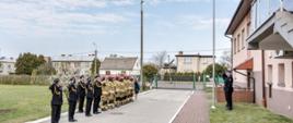 Na asfalcie Przed budynkiem stoją w dwóch rzędach strażacy w ciemnych granatowych i żółtych mundurach. Przed nimi na maszcie powiewa flaga Rzeczypospolitej Polskiej.