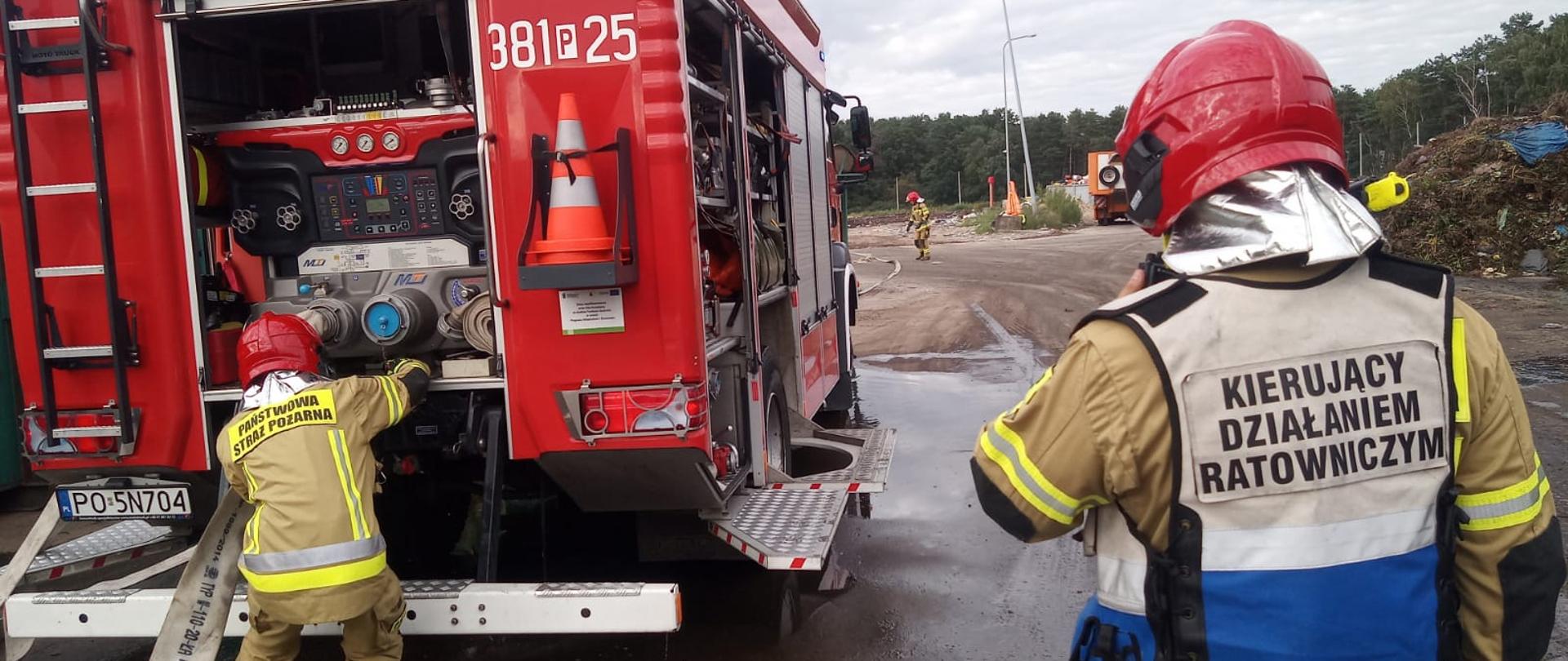 Samochód strażacki stoi na drodze. Strażacy podłączają węże strażackie.
